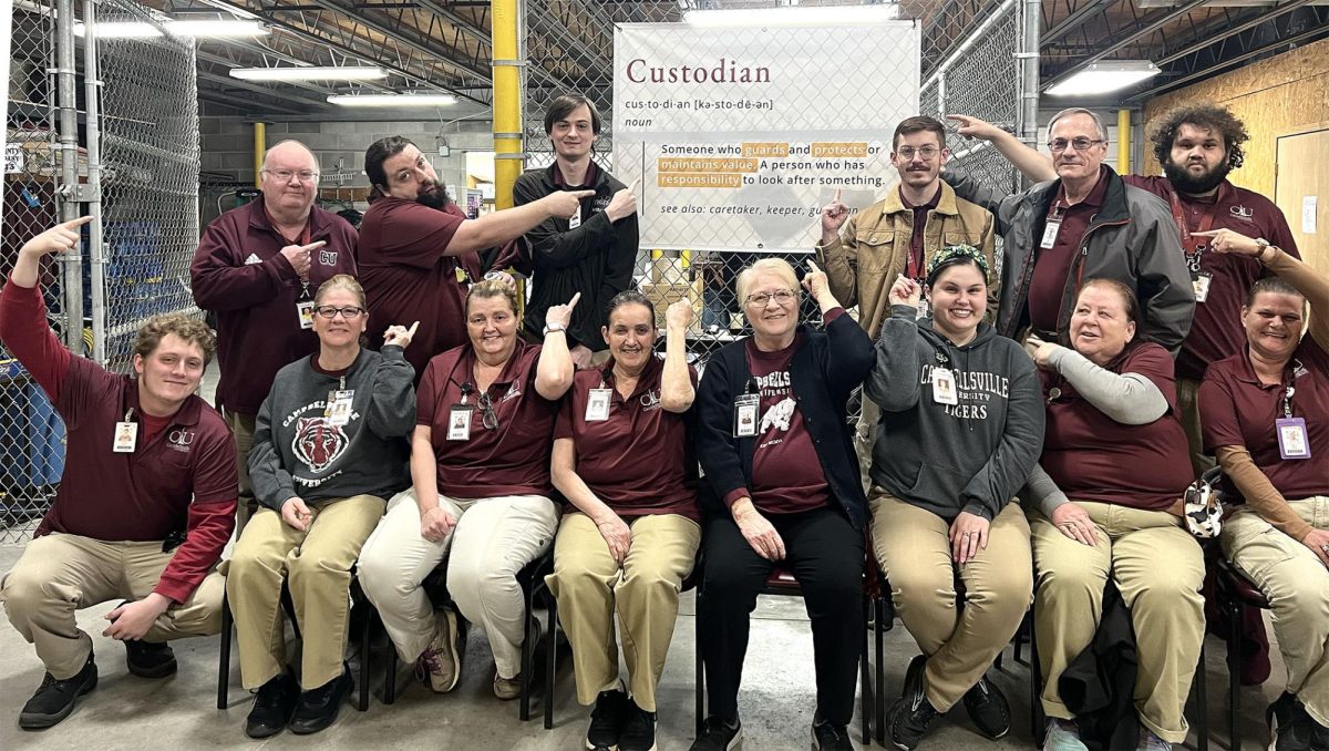 Members of Campbellsville University’s custodial team gather at the physical plant, pointing to a posted definition of “custodian.” Each weekday, the team begins its morning together to pray before heading across campus to clean buildings, fulfill work orders and serve students. Pictured, sitting, from left, are Jordan Proctor, Rhonda Wright, Lorna Bush, Dixie Hourigan, Renee’ Marcum, Hannah Houk, Jackie Abell and Tammy Brown; standing, from left, are James Rice, Zane Wilkins, Brennan Witmer, Caleb Spencer, Scott Iaggi and Dalton Ballentine. (Photo provided)